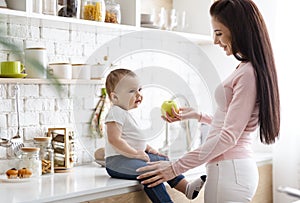 Caring mother offering fresh green apple to her infant at kitchen