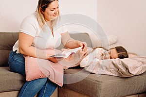 Caring mother reading a book fo her little daughter in the evening while the girl sleeping putting her head on the