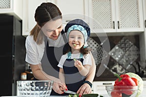 Caring mother teaching little daughter to cook salad in kitchen, young mum and adorable cute girl child wearing apron