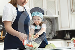 Caring mother teaching little daughter to cook salad in kitchen, young mum and adorable cute girl child wearing apron