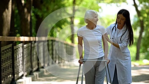 Caring nurse helping senior disabled woman with frame walk in park, rehab photo