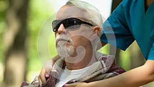 Caring nurse of nursing home covering shoulders of old blind man with plaid