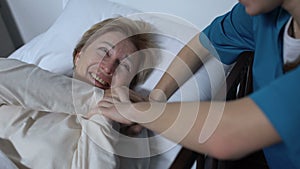 Caring nurse telling jokes to old female patient lying in sickbed rehabilitation