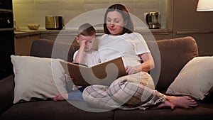 Caring pregnant mother reads book to her beloved blonde child while sitting on sofa in living room in evening. Parent