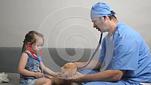 Caring professional male pediatrician playing with small child in room. Little girl plays with a teddy bear Teddy