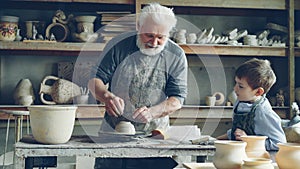 Caring senior grandfather is showing young grandson how to work with clay on throwing-wheel in small workshop. Pottery