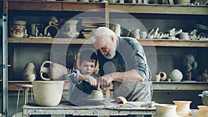 Caring senior grandfather is showing young grandson how to work with clay on throwing-wheel in small workshop. Pottery