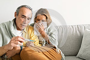 Caring senior man measuring wife's temperature, woman sneezing in napkin, loving husband taking care of ill woman