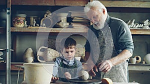 Caring silver-haired grandfather is teaching young cute grandson to work with clay on throwing-wheel in small workshop