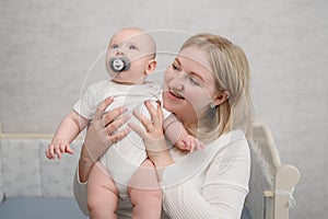 Caring smiling mother holds in her arms, hugs baby with pacifier, taking it from crib, showing love. Portrait of a happy