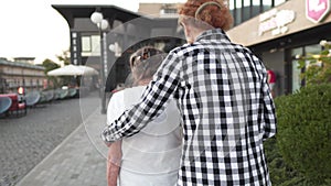 Caring woman with senior citizen walking holding hands and hugging in city. Elderly mom and mature daughter stroll by
