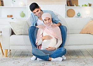 Caring young arab man massaging his wife& x27;s shoulders, resting together at home in living room interior