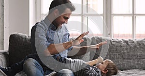 Caring young dad tickling child son laughing playing on sofa
