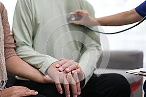 Caring young female doctor examining her contented senior patient with stethoscope in living room. Medical service for elderly,