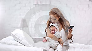 Caring young mother calming down little crying baby during using mobile phone medium shot