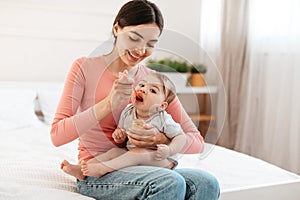 Caring young mother feeding her cute infant baby with mash fruit puree or porridge, giving healthy food to child