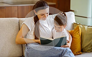 Caring young mother reading book to her cute little daughter at home