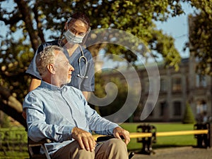Caring young nurse wearing face shield and mask talking to mature man, pushing patient in wheelchair