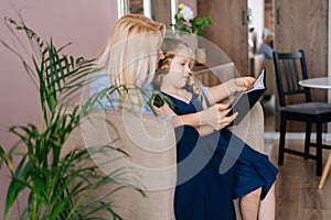 Caring young woman and her little curly daughter reading book together while sitting on armchair