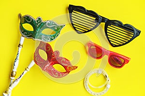 Carnival venetian masks in green and red with handle, fancy glasses and glitter bracelets for photo booth isolated on vibrant yell