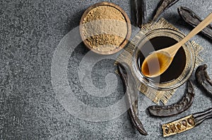 Carob molasses in glass bowl and carob powder or flour with carob pods on rustic background