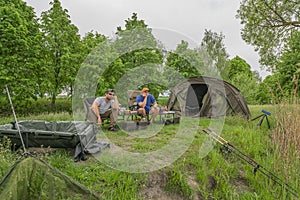 Carp fishing. Two fishermen awaiting for fish bite. Complete equipment and tackle for professional session: carp mat, chairs, tent