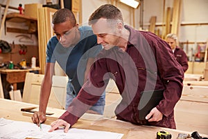 Carpenter With Apprentice Looking At Plans In Workshop