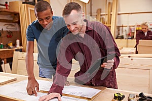 Carpenter With Apprentice Looking At Plans In Workshop