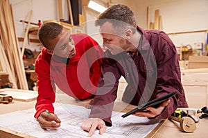 Carpenter With Apprentice Looking At Plans In Workshop