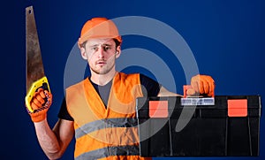 Carpenter concept. Man in helmet, hard hat carries toolbox and holds handsaw, blue background. Worker, repairer
