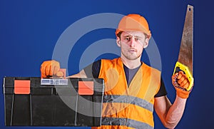 Carpenter concept. Man in helmet, hard hat carries toolbox and holds handsaw, blue background. Worker, repairer