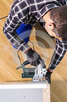 A carpenter with a corded jigsaw cutting a part of the wooden surface (board