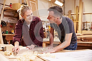 Carpenter With Female Apprentice Planing Wood In Workshop