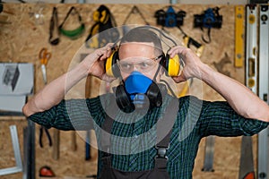A carpenter in goggles and a respirator puts on noise-protection earmuffs.