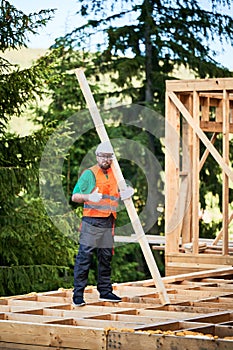 Carpenter holding large beam while builds wooden frame house near the forest.