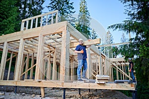 Carpenter holding large beam while builds wooden frame house near the forest.