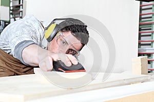 Carpenter man work in the carpentry, sanding wooden boards with sandpaper, protected with ear muffs