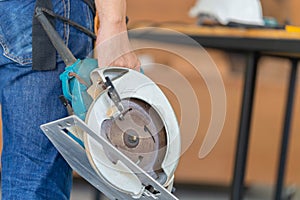 Carpenter man working in wood workshop. Handyman manual worker in tools belt and holding circular saw in his hands