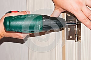 Carpenter mounts wardrobe, screwing a screw, furniture door hinge, using cordless screwdriver.