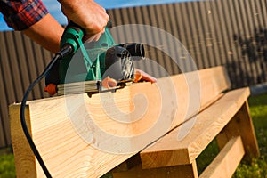 A carpenter planes a wooden board with an electric plane. Planing wooden plank with a electric plane. Close-up on hands with an