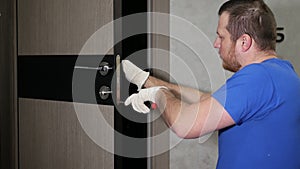 A carpenter in a special uniform repairs the lock of the front door. Repair of the door lock in the apartment. A craftsman repairs