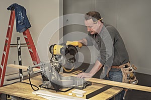 Carpenter with tool belt cutting wood boards with electric chop saw at saw horses during home remodel construction