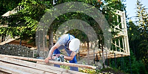 Carpenter using circular saw for cutting wooden plank while building wooden frame house.