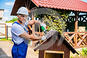 Carpenter using electric screwdriver. Man builds a roof of wooden planks of a small house. Building dog booth