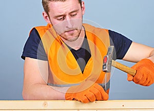 Carpenter, woodworker on concentrated face hammering nail into wooden board. Man, handyman in bright vest and protective