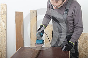 Carpenter working with  electric sheet finishing sander  on wood lath on desk, on background oriented strand board