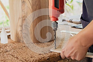 A carpenter is working. He uses an electric drill to drill wood boxes in his small factory