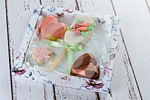 Carrot cake cakesicle surrounded by heart-shaped chocolate and flower-shaped biscuit. On a white box