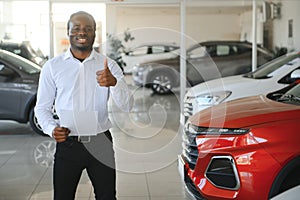 Cars Dealership Concept. Auto Seller Afro Man Standing In Automobile Center