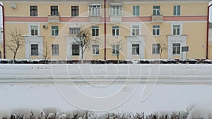 Cars drive along street with old three-storey residential building in winter during snowfall. Roadway is covered with